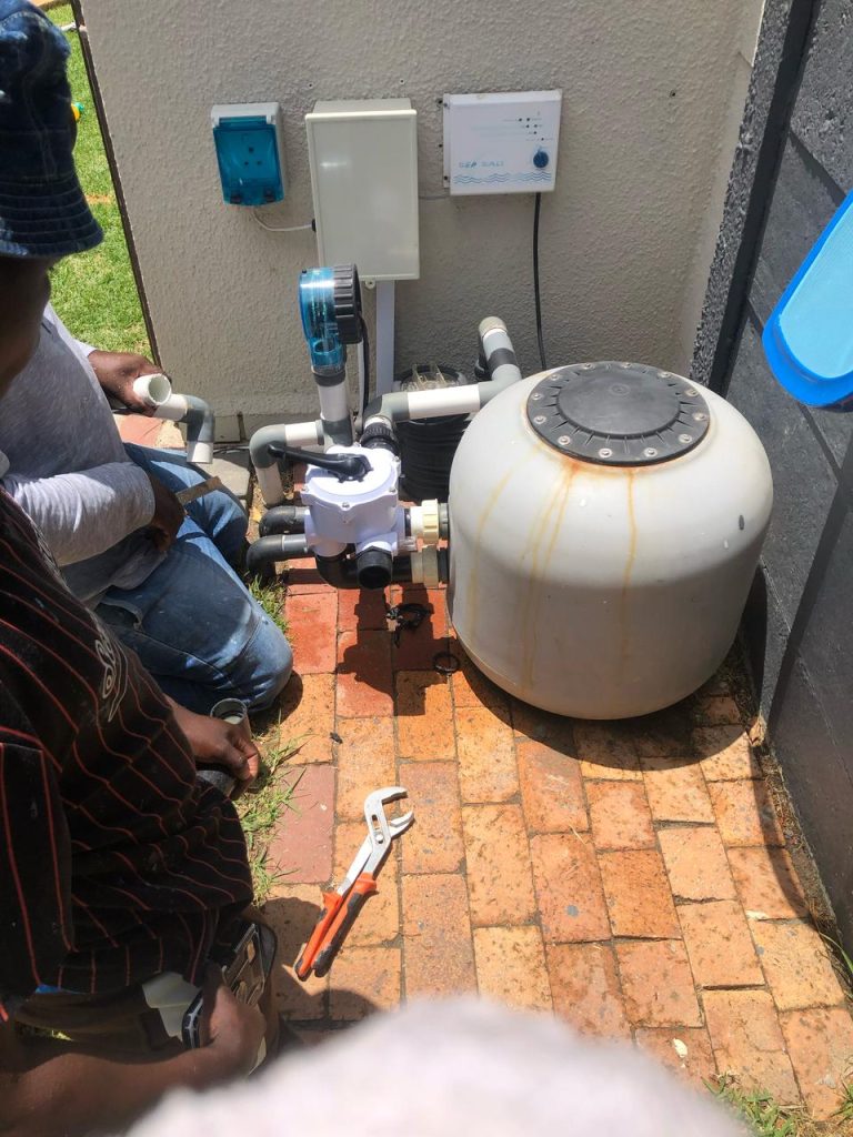 Pool technicians working on the pump and filter plumbing with a pipe wrench beside a sand filter